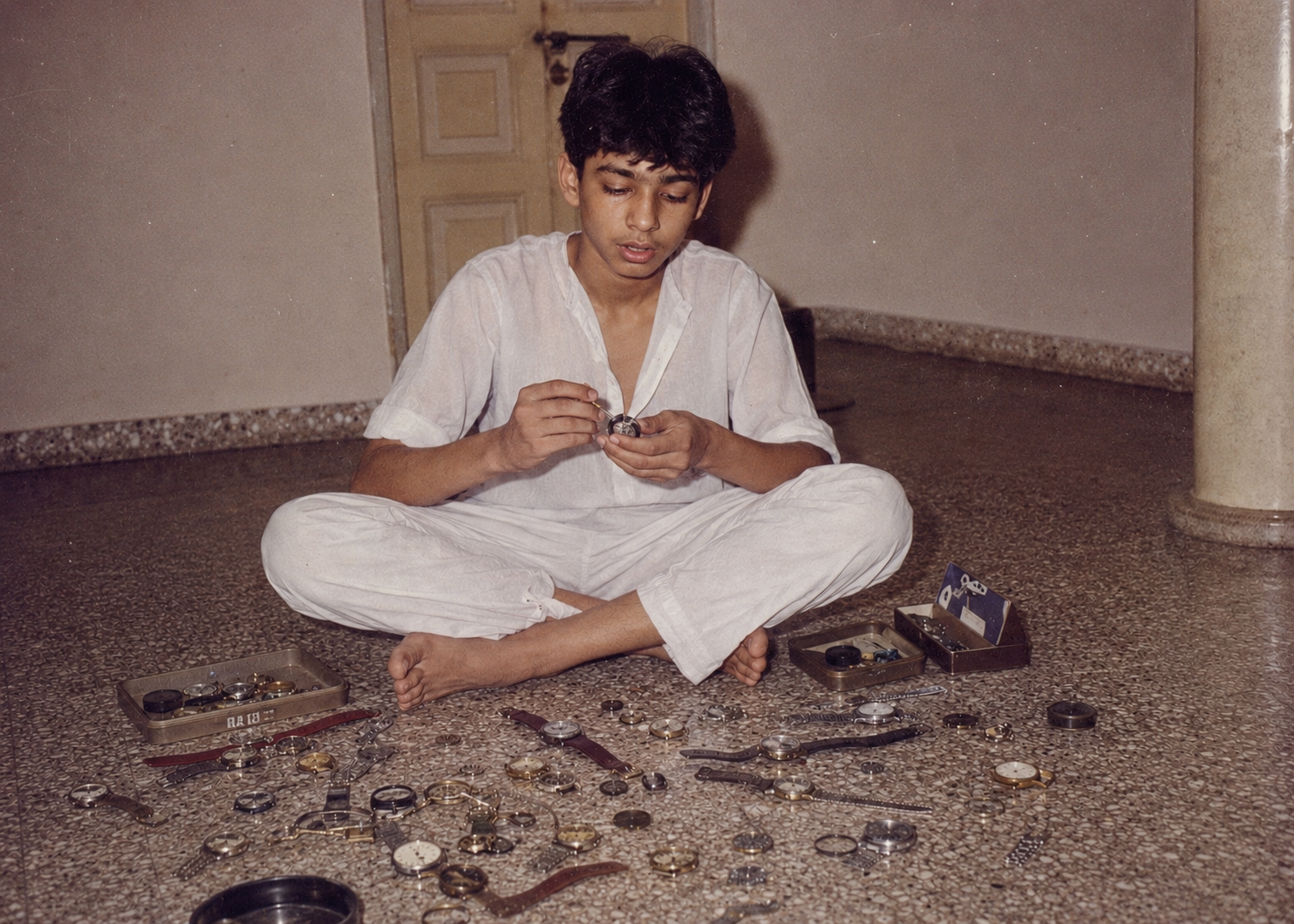 A young Siddhartha seated on the floor, surrounded by opened watches and watch parts